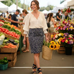 Woman shopping at an outdoor farmers market with produce and flowers around her.