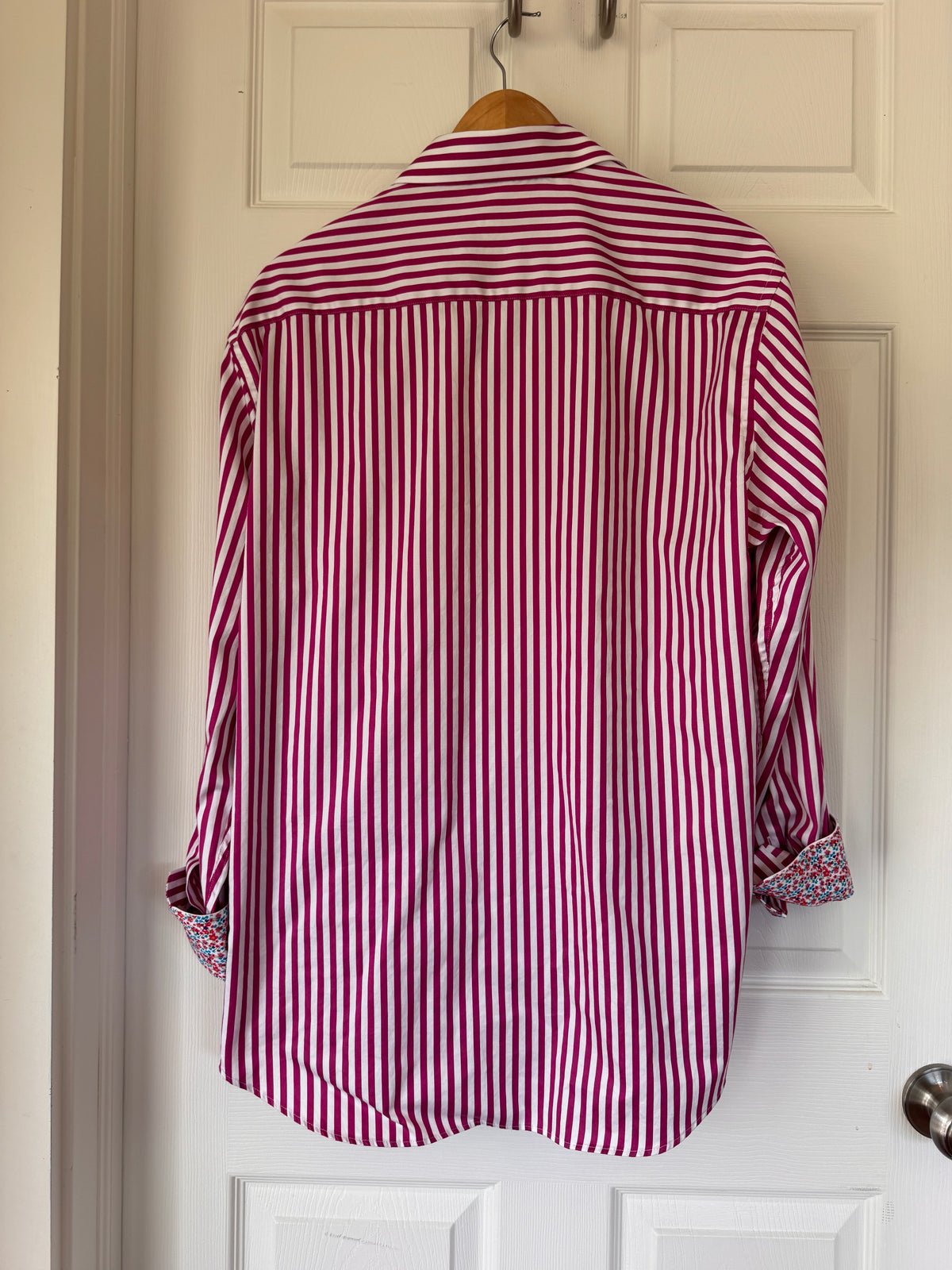 A long-sleeve button-up shirt with vertical red and white stripes, hanging on a wooden hanger against a white paneled door.
