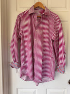 A pink and white vertically striped long-sleeve button-up shirt with multicolor floral patterned cuffs, hanging on a wooden hanger against a white paneled door.