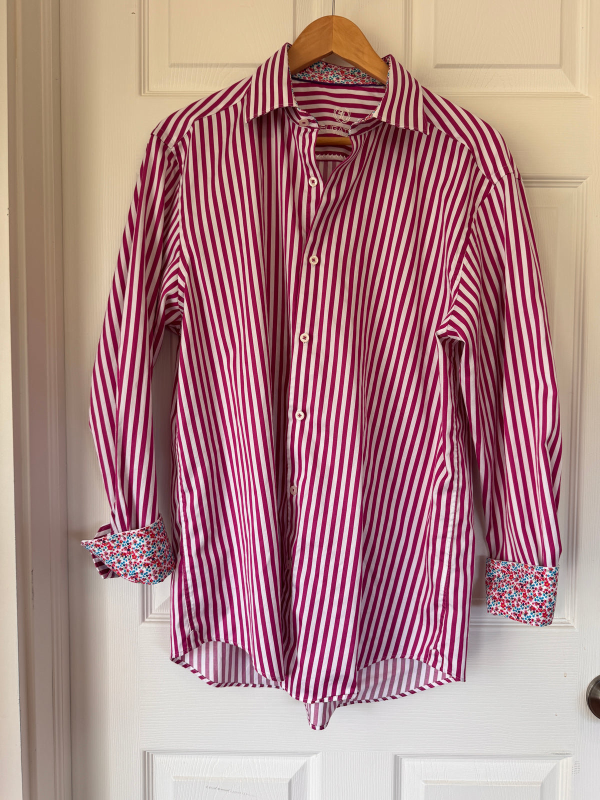 A pink and white vertically striped long-sleeve button-up shirt with multicolor floral patterned cuffs, hanging on a wooden hanger against a white paneled door.