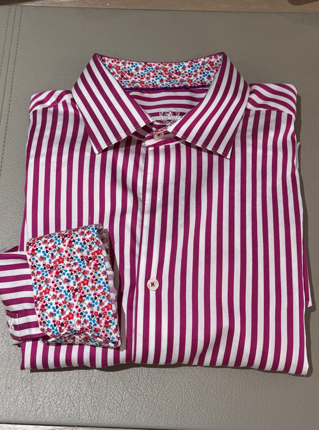 A folded long-sleeve dress shirt with vertical magenta and white stripes, featuring a contrasting multicolor floral pattern on the inner collar and cuff, displayed on a gray textured surface.