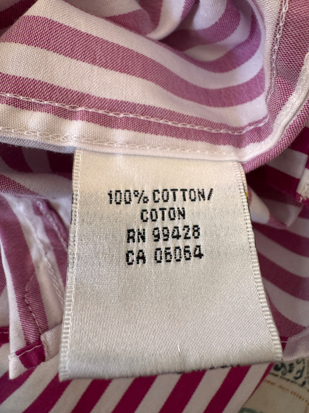 Close-up of a garment's interior neck label reading '100% COTTON' with pink and white striped fabric visible around it.
