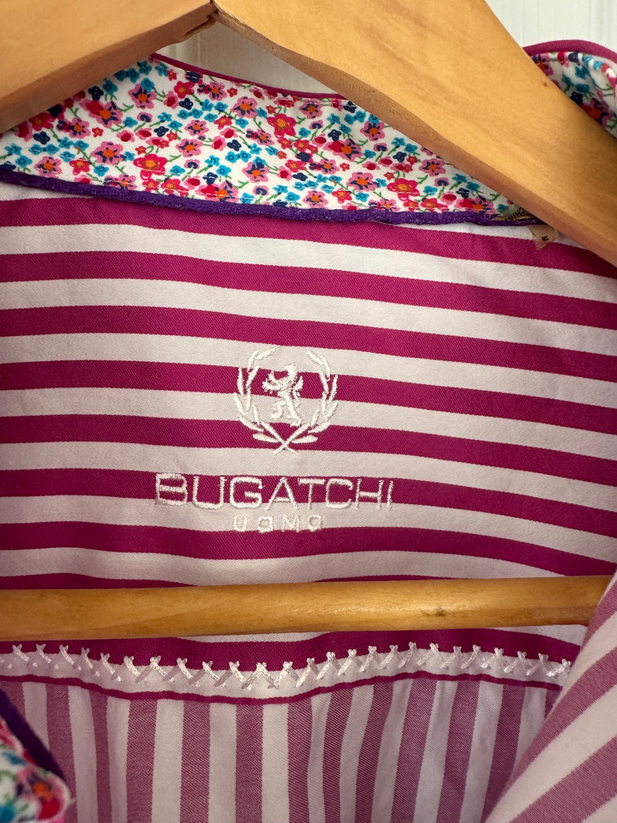 A close-up of a striped garment hanging on a wooden hanger, featuring magenta and white horizontal stripes with a white embroidered Bugatchi UOMO logo in the center. The top edge shows a colorful floral pattern lining.