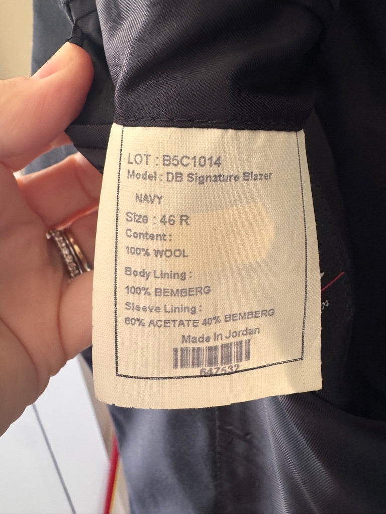 A close-up of a garment's interior label showing details for the 'DB Signature Blazer' in navy wool, held by a hand with a ring.