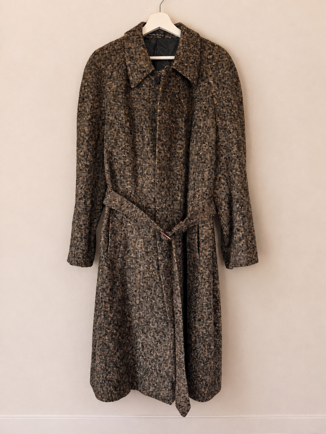 A long-sleeved, knee-length coat with a matching waist tie belt, featuring a textured mottled pattern of brown, black, and beige. It is shown hanging on a white hanger against a plain, warm beige wall.