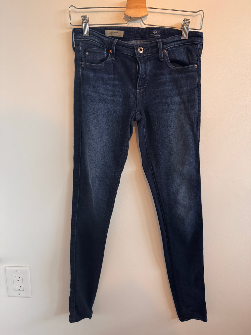 Pair of dark blue jeans on a wooden hanger against a beige wall.