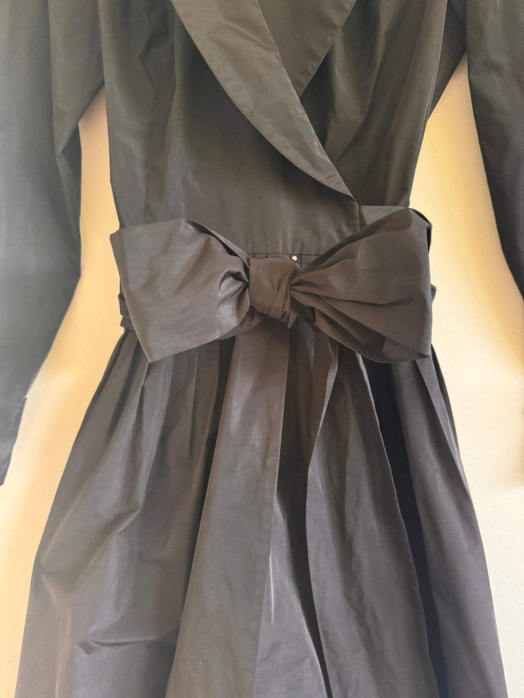 A close-up of a black wrap-style dress or coat with a large bow belt at the waist, shown against a plain light background.