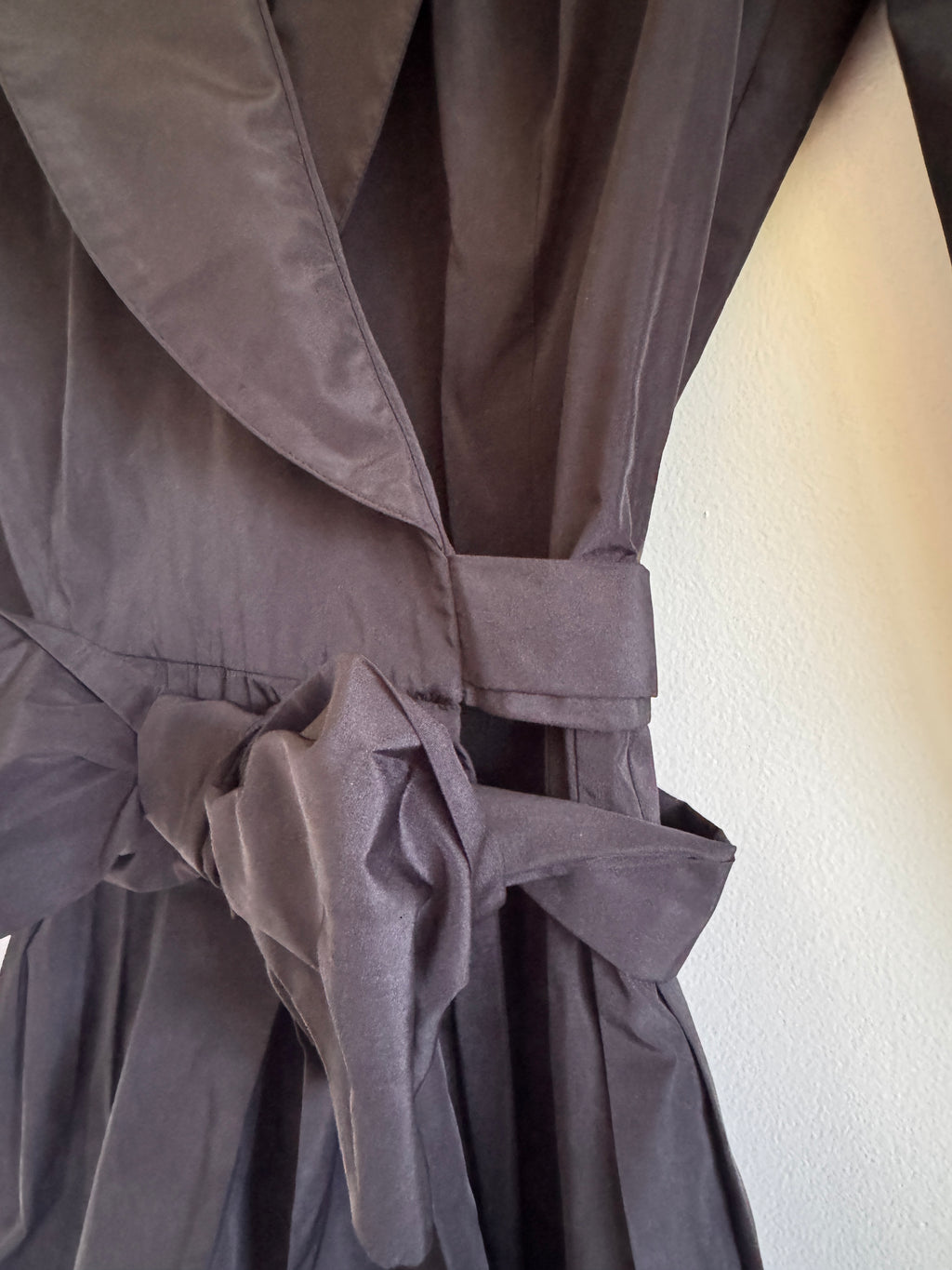Close-up of a black fabric garment with gathered pleats and a tie detail, photographed against a light wall.