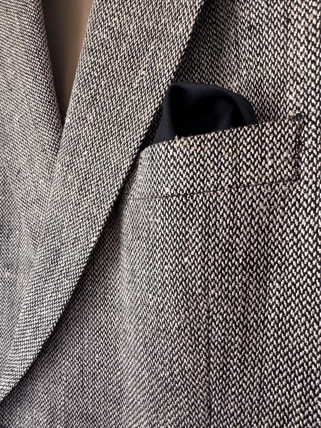 Close-up of a textured herringbone tweed blazer in gray and beige, with a black pocket square tucked into the chest pocket.