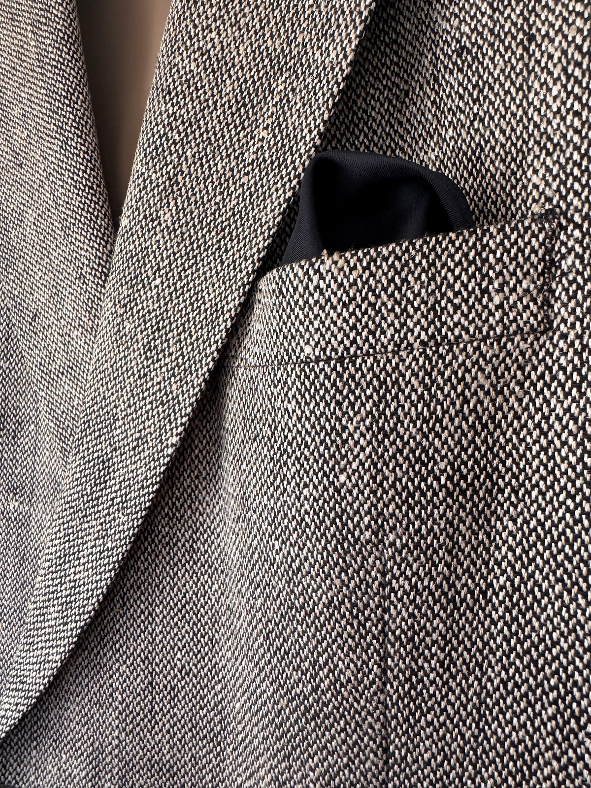 Close-up of a textured herringbone tweed blazer in gray and beige, with a black pocket square tucked into the chest pocket.