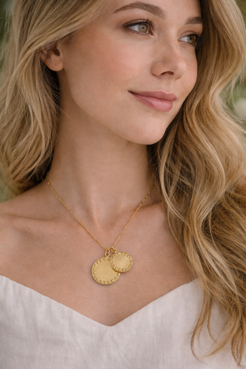 Woman wearing a gold necklace with two round pendants, smiling outdoors.