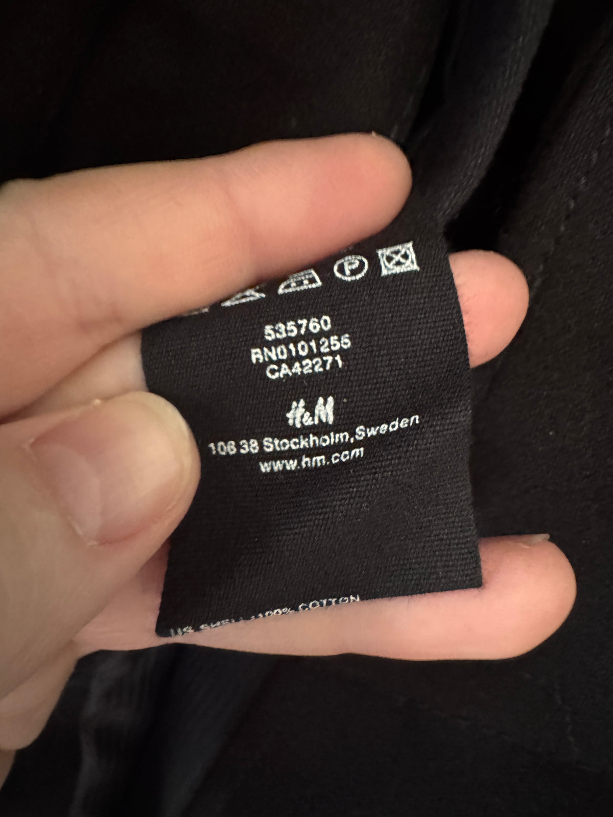 A close-up of a hand holding a black fabric clothing tag with white printed text and symbols. The tag shows the H&M logo and Stockholm, Sweden address.