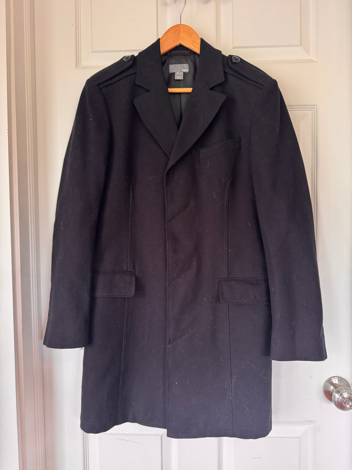 A black wool-blend military-style overcoat hanging on a wooden hanger against a white paneled door.