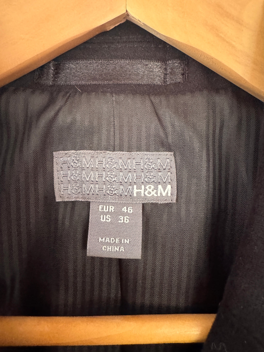 Close-up of the interior neck label of a black H&M garment hanging on a wooden hanger, showing the H&M brand name and size information.