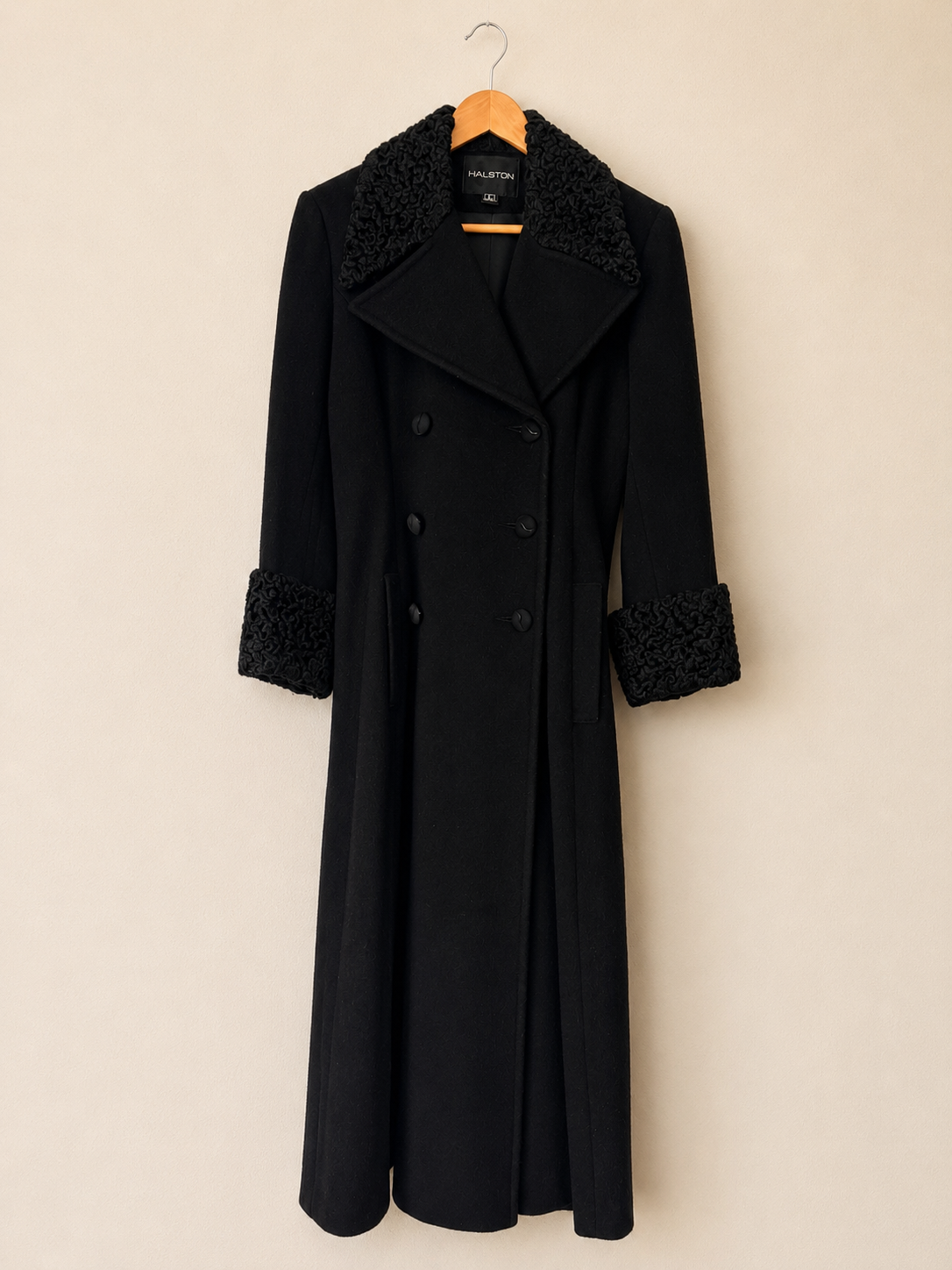 A long black double-breasted coat with textured black lace trim on the collar, cuffs, and button placket, hanging on a wooden hanger against a plain beige wall.