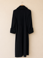 A long black coat hanging on a wooden hanger against a plain beige wall. The coat features a textured black knit collar and matching textured trim at the cuffs.
