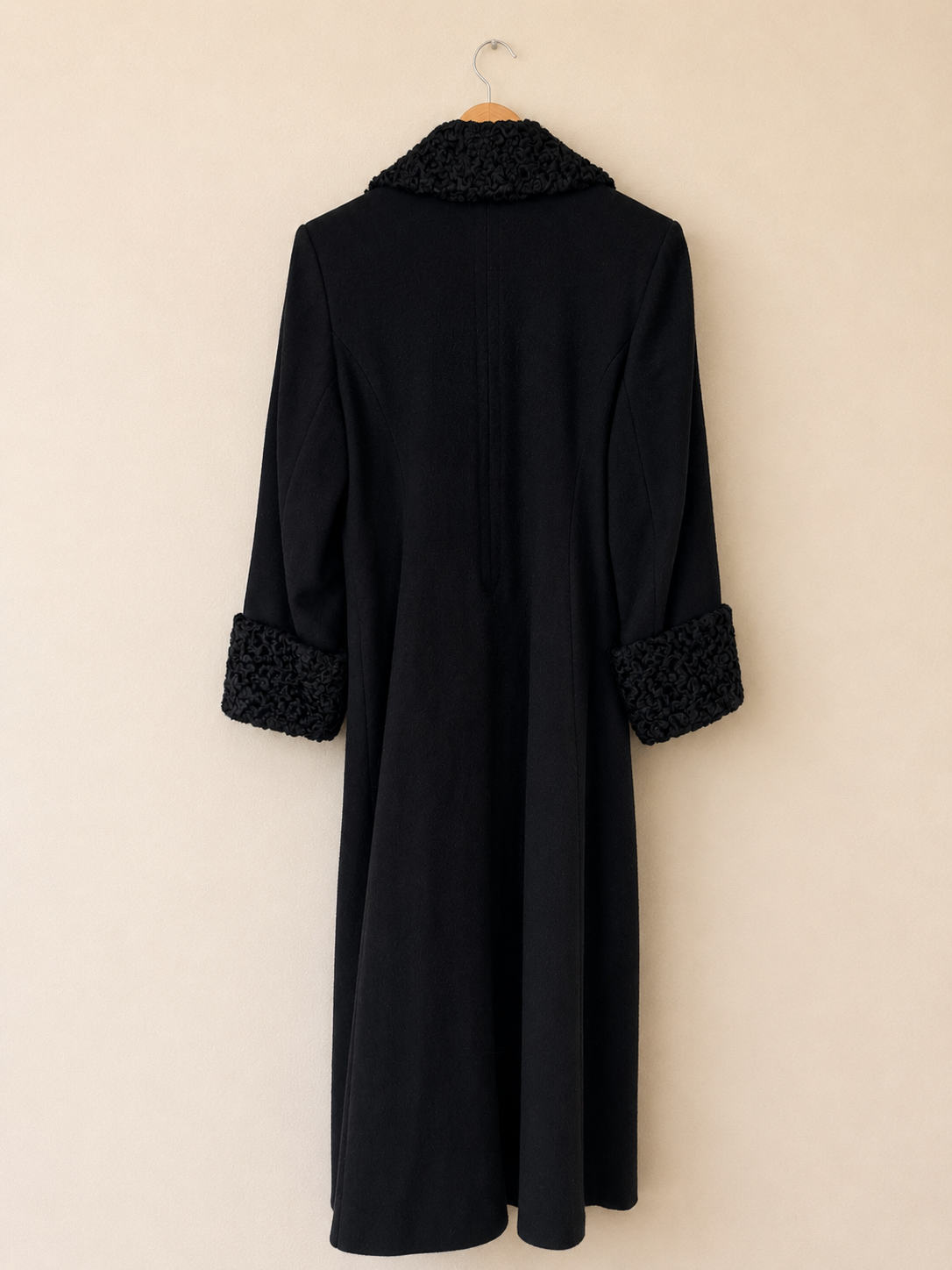 A long black coat hanging on a wooden hanger against a plain beige wall. The coat features a textured black knit collar and matching textured trim at the cuffs.