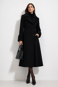 A woman models a long black double-breasted coat with textured black fur trim on the collar and cuffs, paired with a black turtleneck, black tights, black heels, and a black handbag against a plain white studio background.