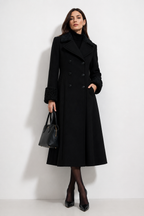A woman models a long black double-breasted coat with textured black fur trim on the collar and cuffs, paired with a black turtleneck, black tights, black heels, and a black handbag against a plain white studio background.