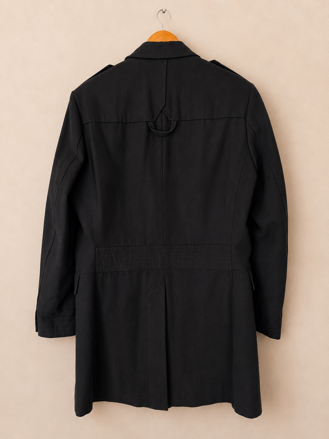 A black longline trench coat with a belted waist and shoulder epaulets, hanging on a wooden hanger against a solid beige wall.