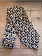 A men's necktie with a repeating Art Deco geometric pattern in black, gold, and light blue, laid flat on a rustic wooden surface.