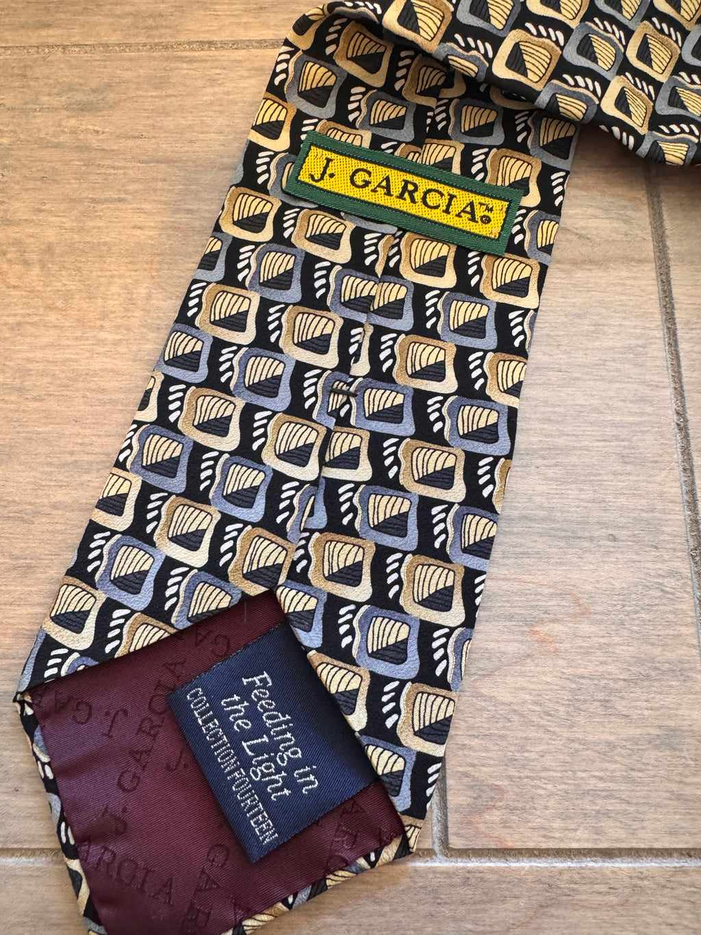 A J. Garcia men's necktie with a geometric pattern in black, gold, and gray, laid flat on a light wood background.