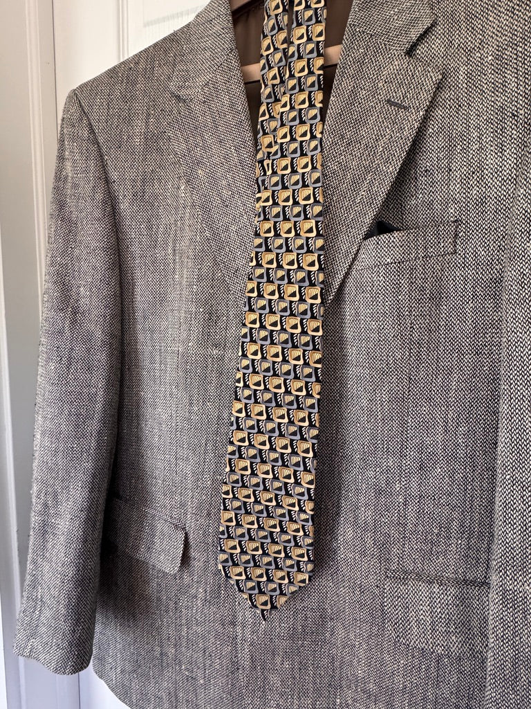A men's gray textured herringbone blazer jacket displayed on a hanger with a patterned necktie and pocket square.