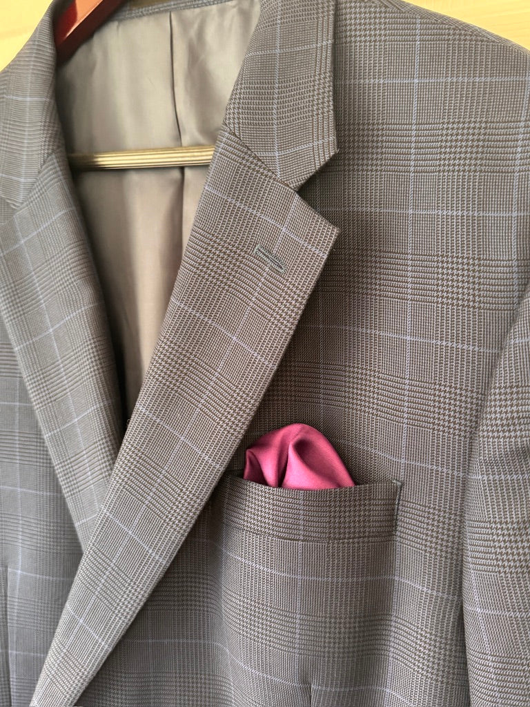 A close-up of a blue and black plaid blazer jacket with a pink pocket square tucked into the chest pocket, hanging on a wooden hanger.
