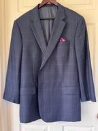 A navy blue windowpane plaid blazer with a pink pocket square, hanging on a wooden hanger against a cream-colored paneled door.