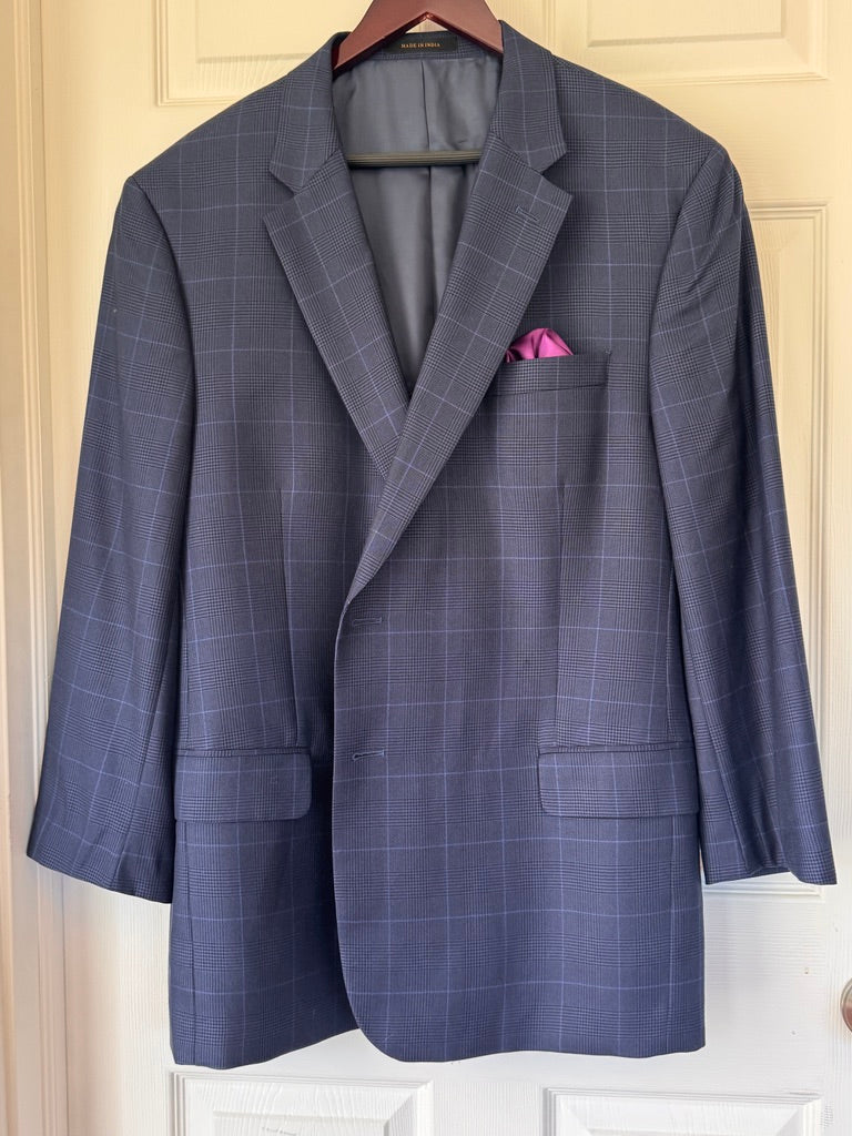 A navy blue windowpane plaid blazer with a pink pocket square, hanging on a wooden hanger against a cream-colored paneled door.