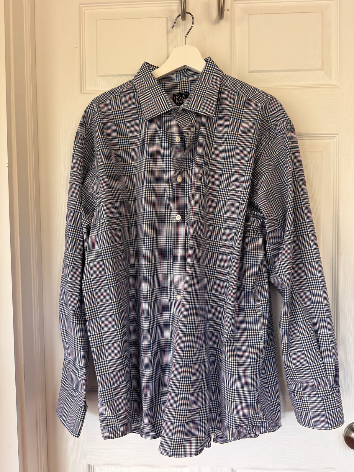 A long-sleeve plaid button-up shirt in blue, white, and red, hanging on a white hanger against a white paneled door.