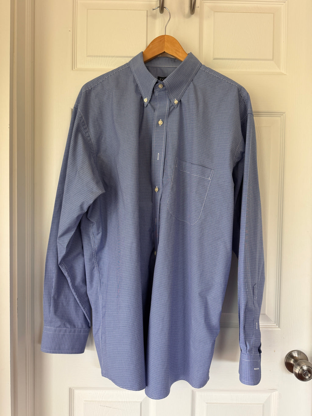 A blue long-sleeve button-down shirt with a subtle grid pattern, hanging on a wooden hanger against an off-white paneled door.