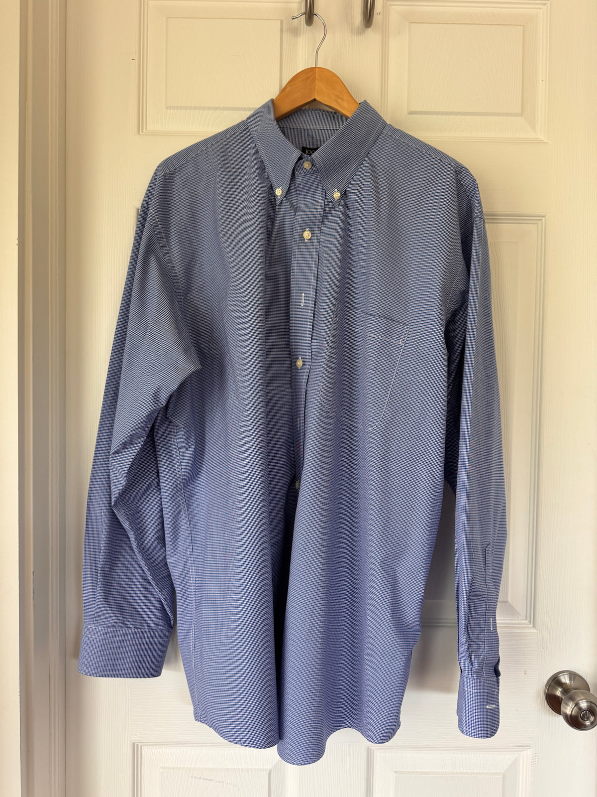A blue long-sleeve button-down shirt with a subtle grid pattern, hanging on a wooden hanger against an off-white paneled door.