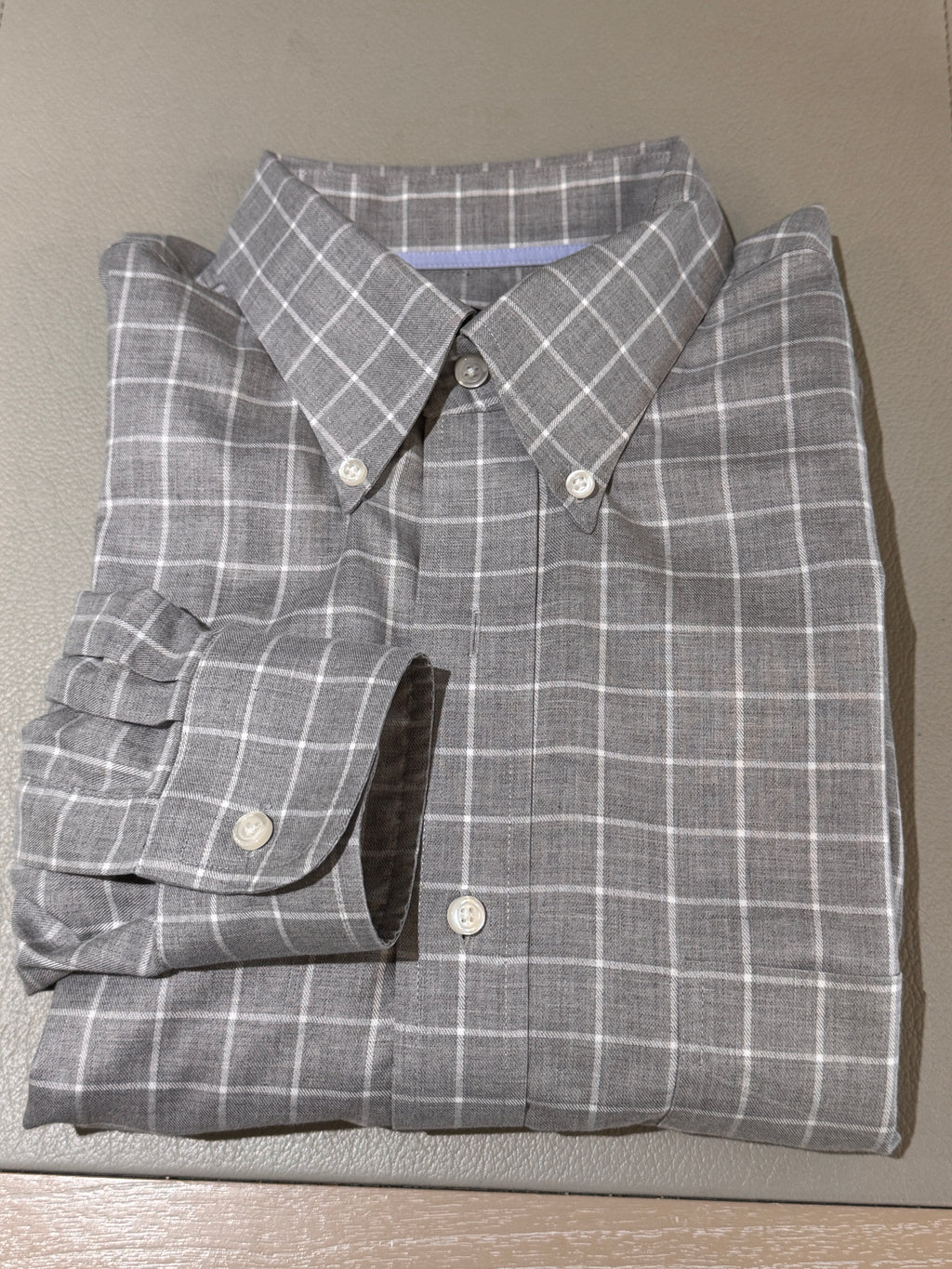 A folded gray plaid button-down shirt with white grid lines, laid flat on a textured beige surface.