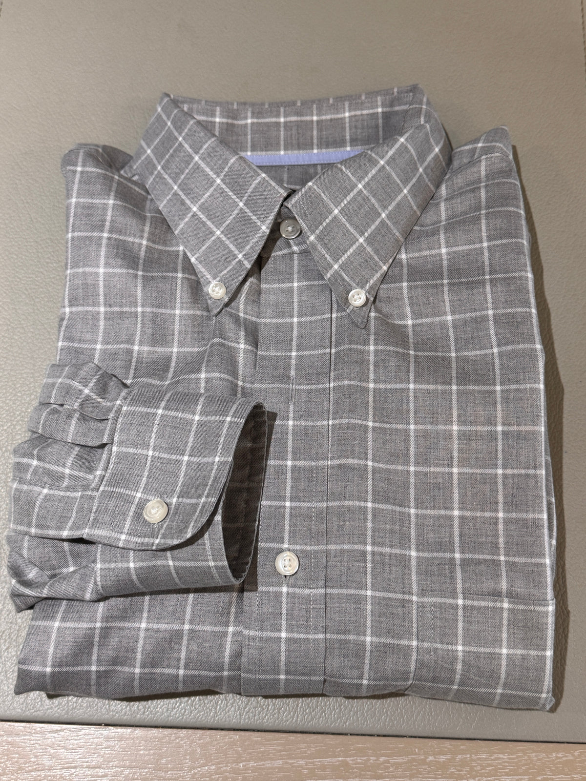 A folded gray plaid button-down shirt with white grid lines, laid flat on a textured beige surface.