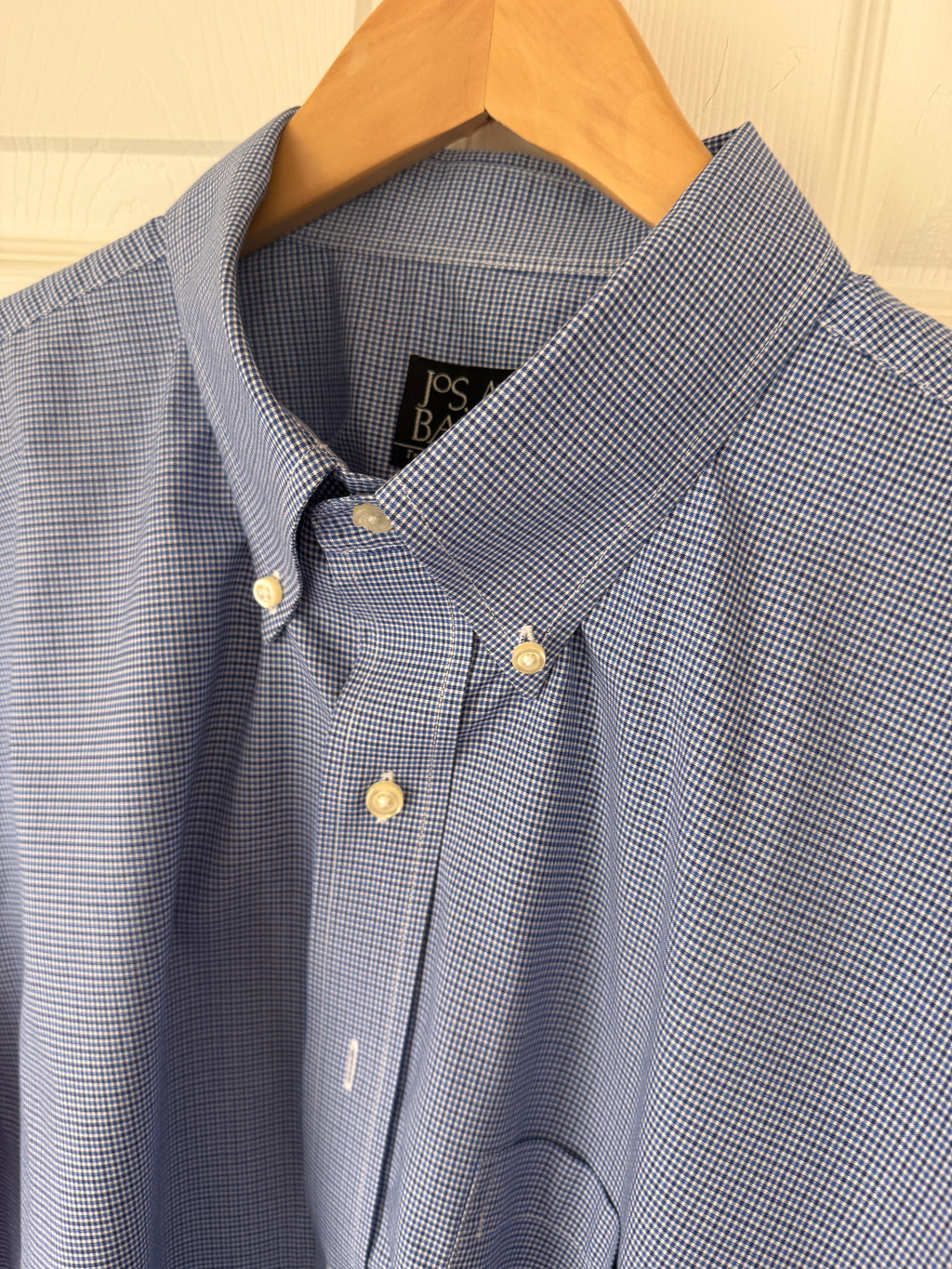 A close-up of a blue and white micro-check patterned button-down shirt hanging on a wooden hanger against a light-colored paneled door.
