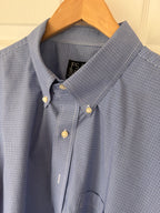 A close-up of a blue and white micro-check patterned button-down shirt hanging on a wooden hanger against a light-colored paneled door.