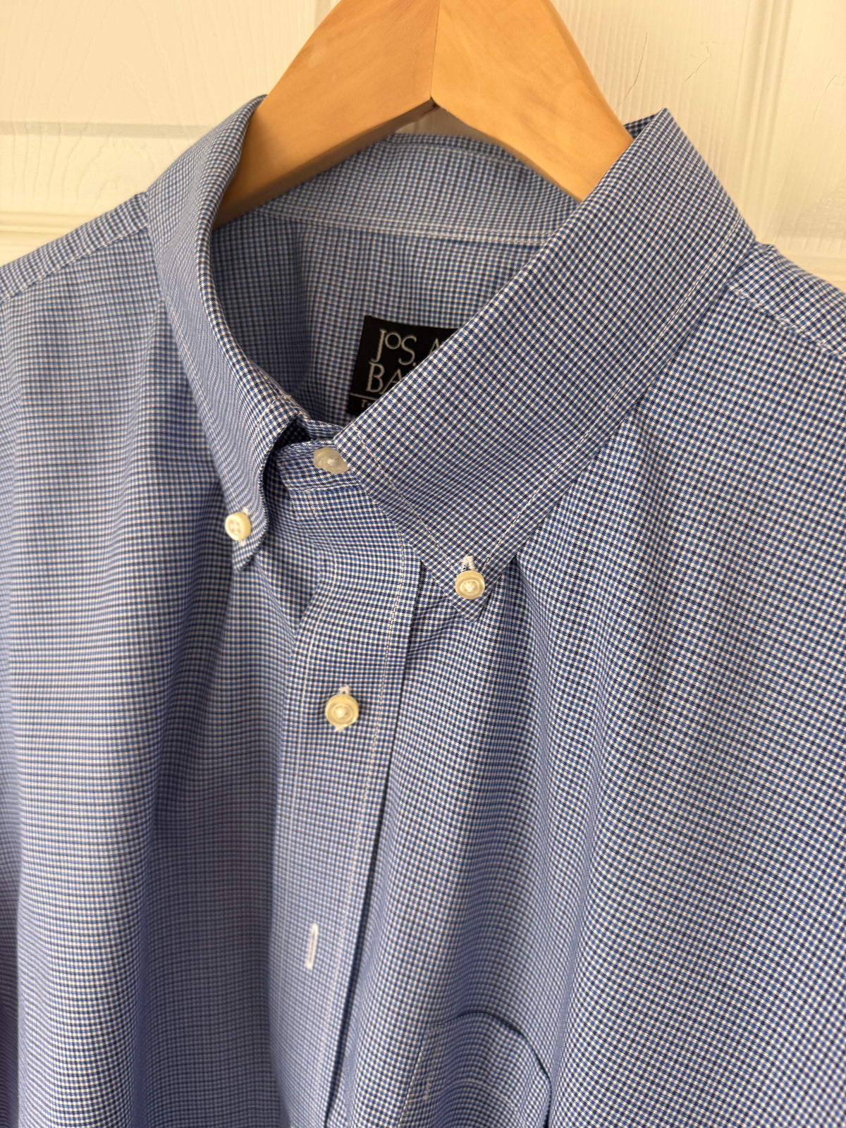 A close-up of a blue and white micro-check patterned button-down shirt hanging on a wooden hanger against a light-colored paneled door.