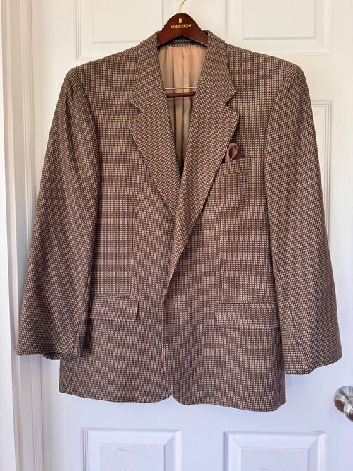 A brown and beige houndstooth patterned blazer jacket hanging on a wooden hanger against a white paneled door.