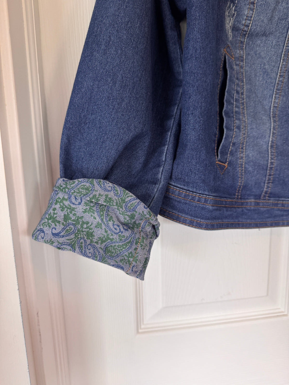 A close-up of a blue denim jacket hanging on a white door, showing a rolled-up sleeve with a green and blue paisley patterned lining.