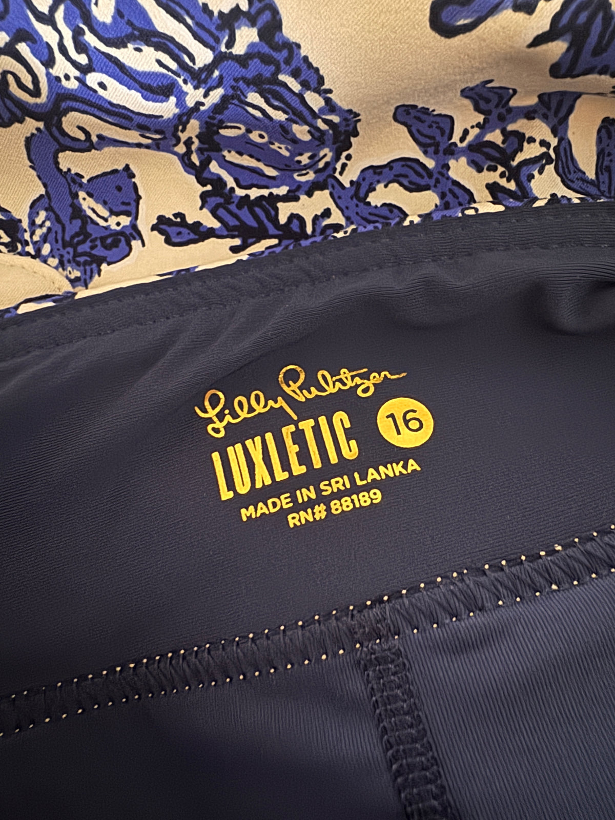 Close-up of a navy blue garment interior label reading 'Lil' Luxton LUXLETIC 16 MADE IN SRI LANKA RN# 88189' on dark fabric, with a beige and blue patterned section visible above.
