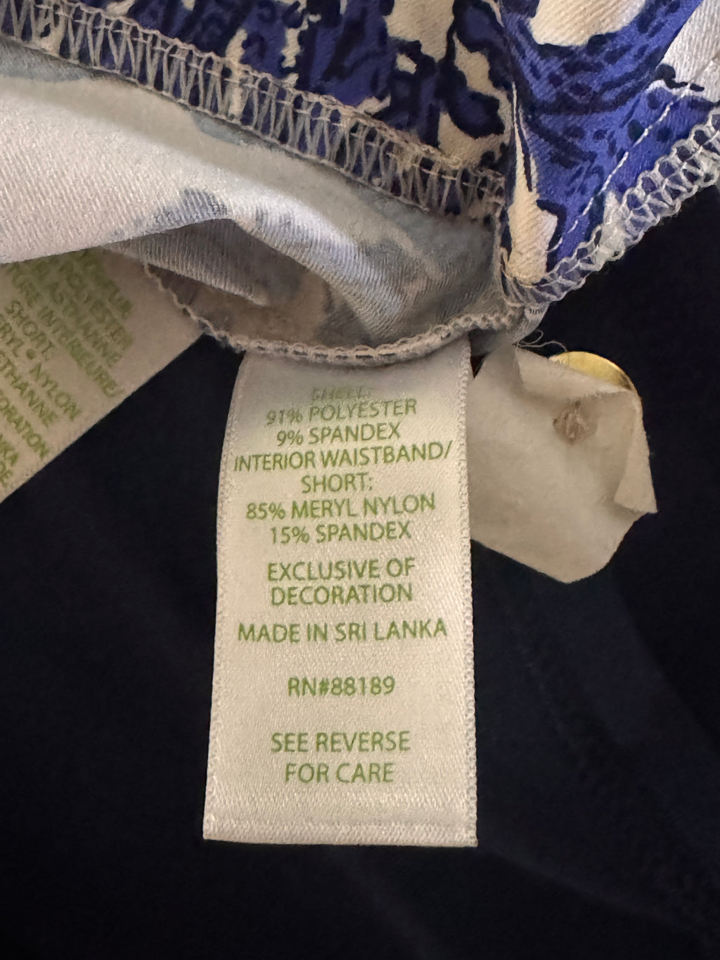 Close-up of a garment's interior waistband and care label, showing a white fabric tag with green text reading 'COTTON 95% POLYESTER 9% SPANDEX INTERIOR WAISTBAND/SHORT: 85% MERYL NYLON 15% SPANDEX EXCLUSIVE OF DECORATION MADE IN SRI LANKA RN#88189 SEE REVERSE FOR CARE.'.