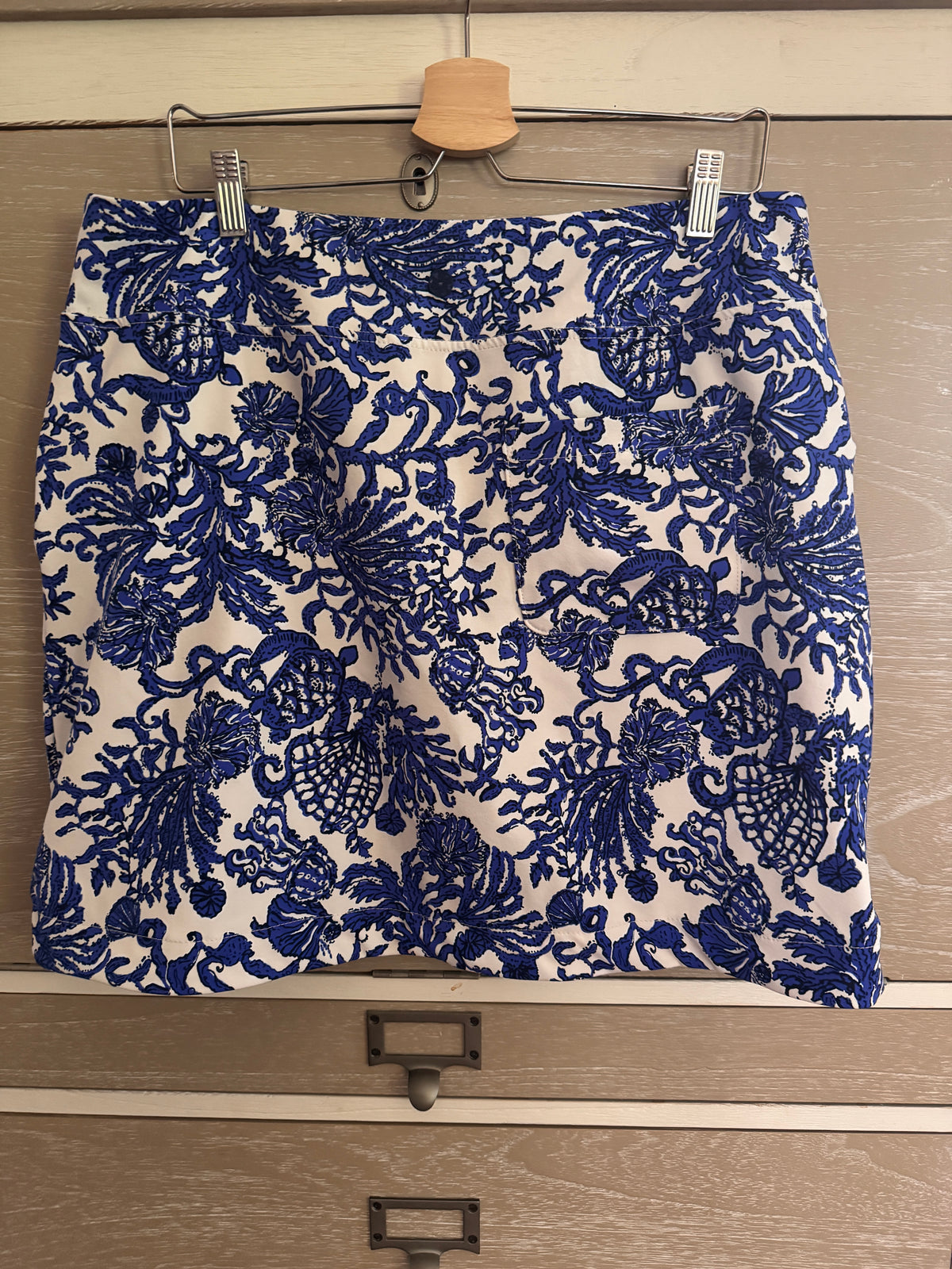 A blue and white jacquard patterned skirt hanging from a wooden hanger on a light wood drawer front.