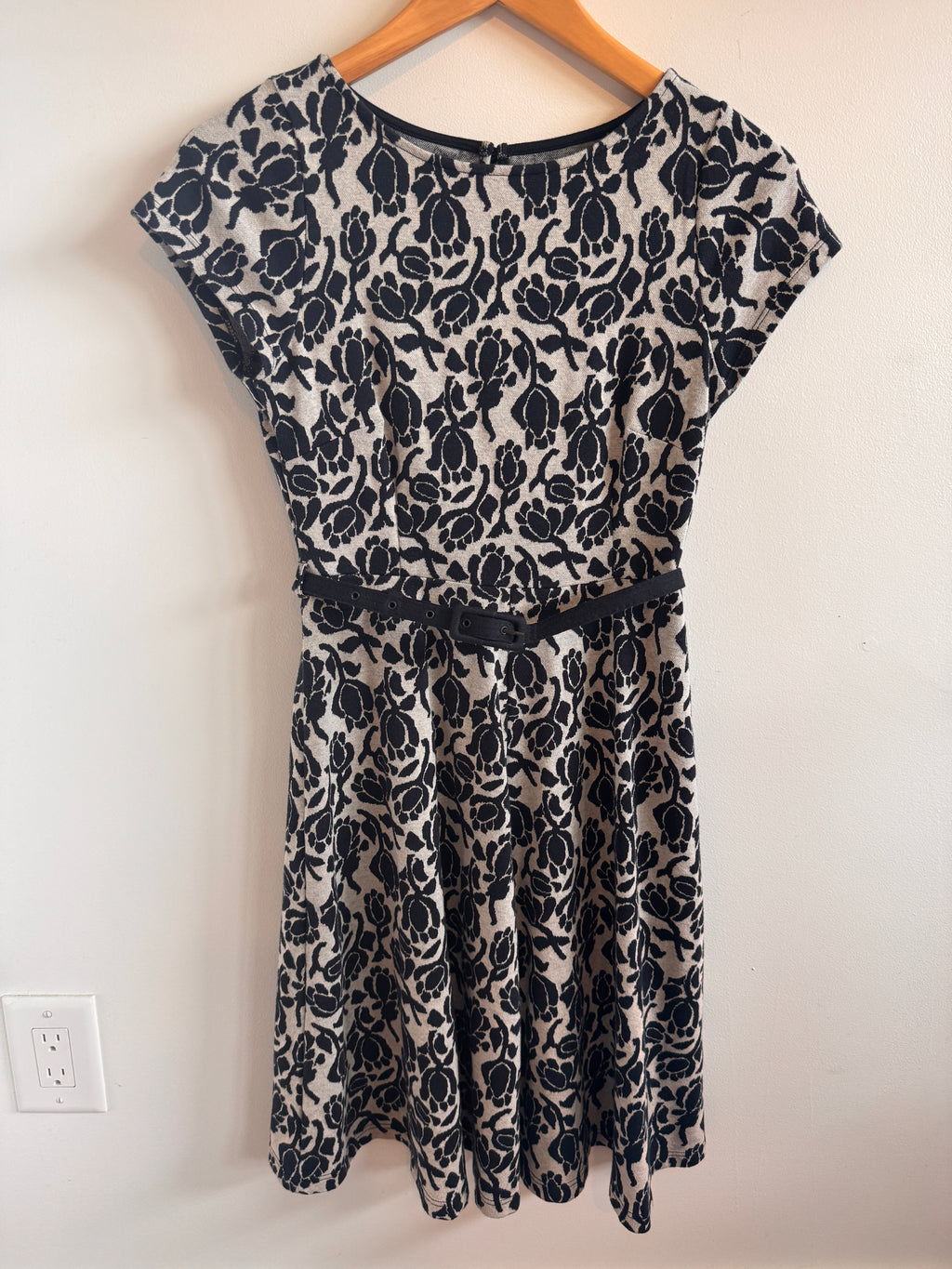 Black floral-patterned dress on a wooden hanger against a white wall.