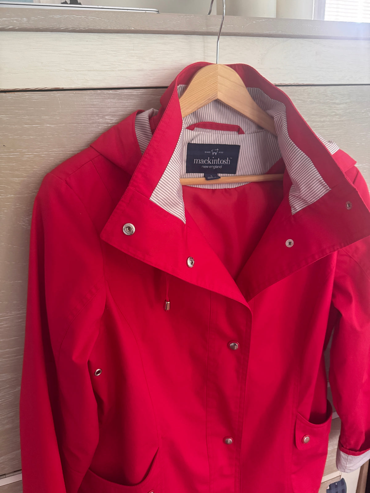 A bright red hooded raincoat from the brand Mocktailosh hanging on a wooden hanger against a light wood-paneled wall.