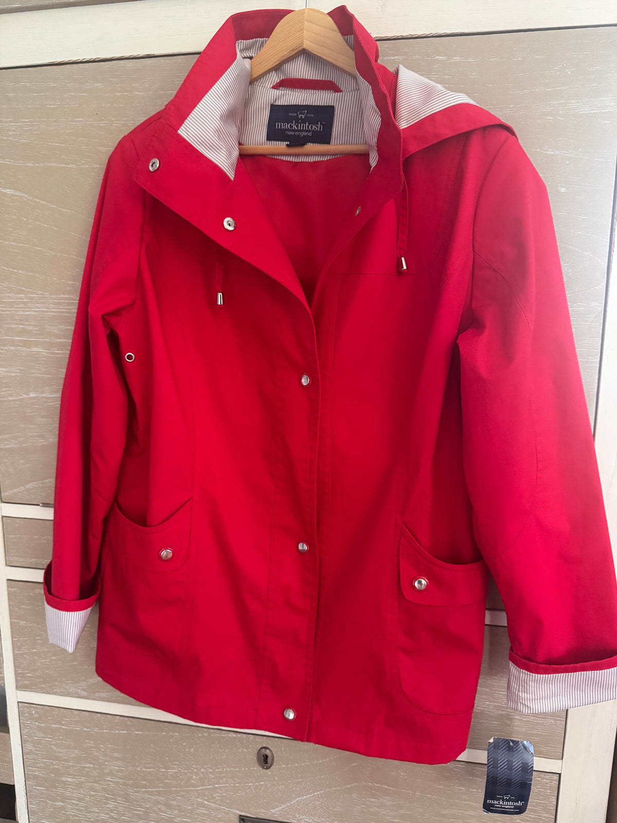 A bright red hooded raincoat hanging on a wooden hanger against a light wood-paneled wall. The coat has a white hood lining, silver snap buttons, and front patch pockets.