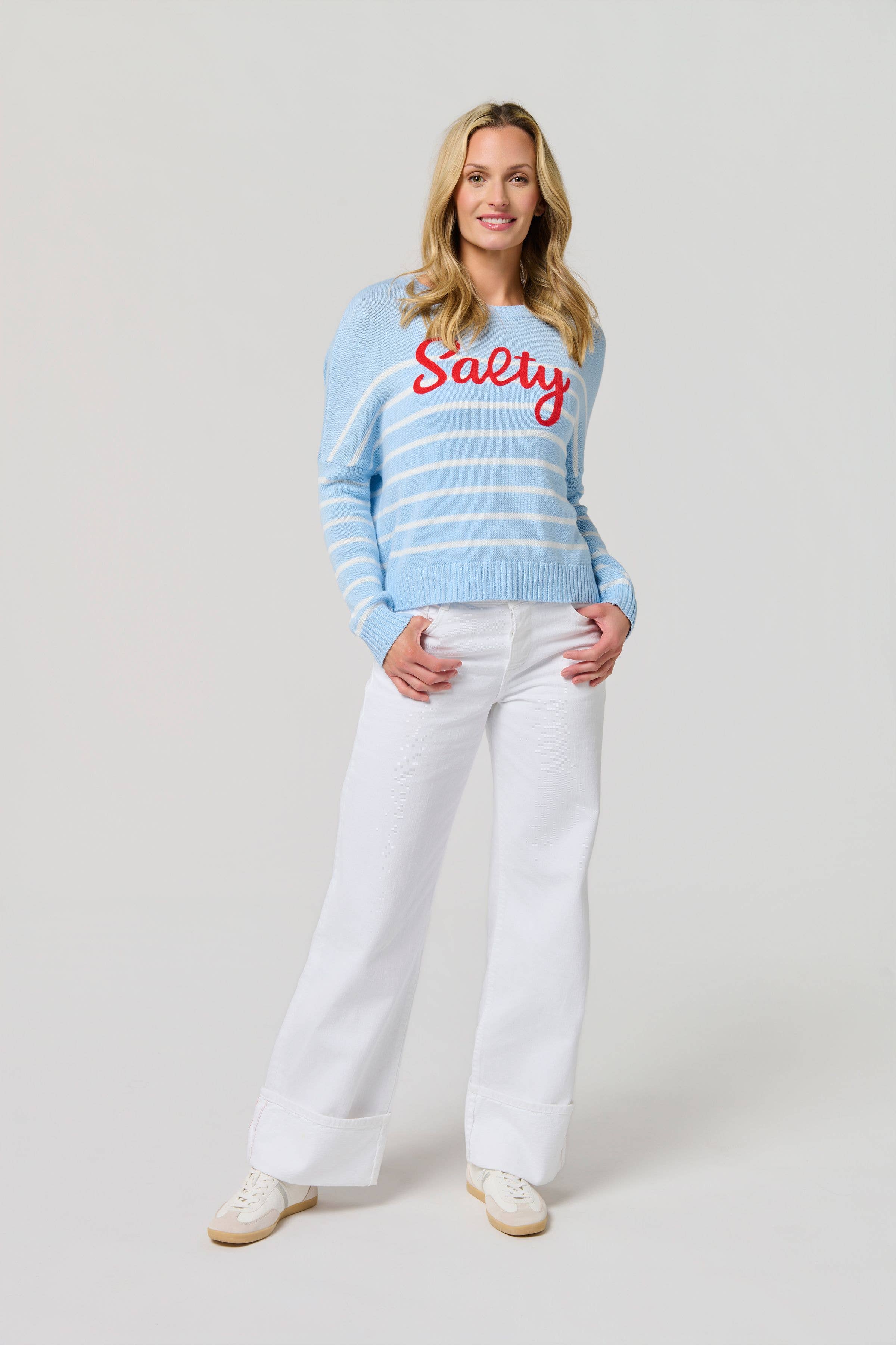 Woman wearing a light blue striped sweater with 'Salty' text and white pants on a plain background