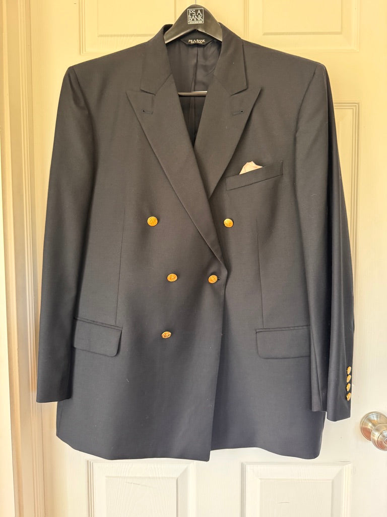A navy double-breasted blazer with gold buttons, hanging on a black hanger against a cream-colored paneled door. It features notched lapels, a chest pocket with a white pocket square, and flap pockets.