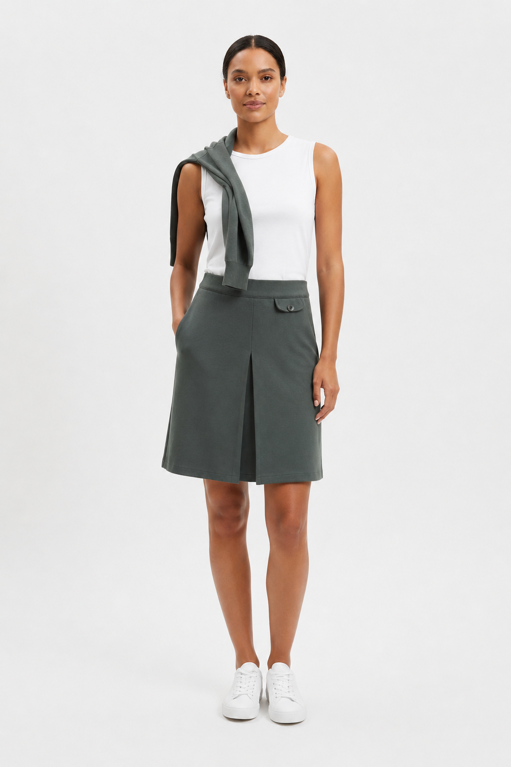 Woman wearing a skirt with a white top and matching sweater over her shoulder against a white background