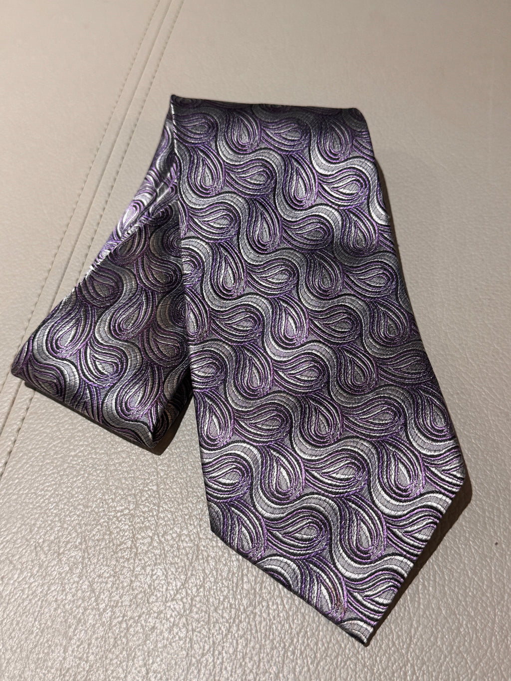 A patterned necktie with a purple, gray, and white wavy paisley design, laid flat on a light beige textured surface.